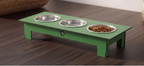A green elevated pet feeder and water bowl stand with three stainless steel bowls, one filled with dry kibble, shown on a dark wood floor.