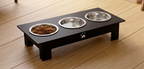 A black elevated pet feeder stand with three stainless steel bowls, shown on a light wood floor. One bowl contains dry dog food.