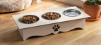 A white elevated pet feeder and water bowl stand on a wooden floor, with two metal bowls containing pet food and one empty water bowl. The stand has a black paw print cutout on the front.