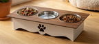 An elevated pet feeder and water bowl stand with a wood-grain top, white base, and black paw print cutout, shown on a wooden floor with food and water bowls filled.
