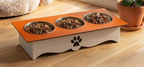 A three-compartment elevated pet feeder with a white base, orange top panel, and stainless steel bowls filled with wet food, placed on a wooden floor in a home setting.
