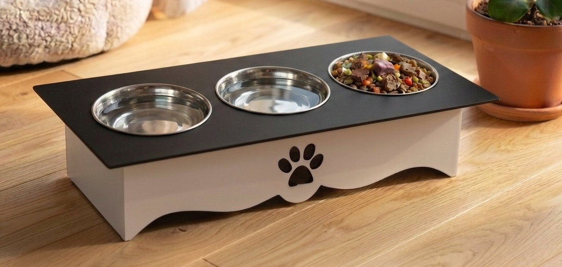An elevated pet feeder and waterer stand with three bowls, shown on a light wood floor in a home setting. The stand has a white base with a black paw print cutout, a black top tray, stainless steel bowls, and a bowl of wet food on the right.
