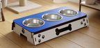 A three-bowl elevated pet feeder and water with a blue top surface, stainless steel bowls, and a white base decorated with black paw prints and a bone cutout, shown on a wooden floor.
