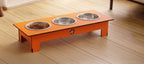 An orange elevated pet feeder stand with three stainless steel bowls, one filled with dry dog food, placed on a light wood floor.