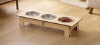 A wooden elevated pet feeder and water bowl stand with three stainless steel bowls, one filled with dry pet food, set on a light wood floor in a home interior.