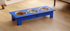 A blue elevated pet feeder stand with three stainless steel bowls, one filled with dry pet food, placed on a light wood floor.