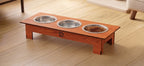 A wooden elevated pet feeder stand with three stainless steel bowls, one filled with dry pet food, placed on a light wood floor in a home setting.