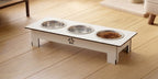 A white elevated pet feeder and water bowl stand with three stainless steel bowls, one filled with dry kibble, placed on a light wood floor.