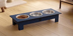 A dark blue elevated pet feeder and water bowl stand with three stainless steel bowls, shown on a light wood floor in a home setting.