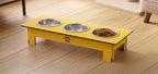 A yellow elevated pet feeder and water bowl stand with three stainless steel bowls, shown on a light wood floor in a home setting.