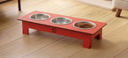 A red elevated pet feeder and water bowl stand with three stainless steel bowls, one filled with dry food, placed on a light wood floor.