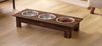 A dark brown wooden elevated pet feeder and water bowl stand with three stainless steel bowls, one containing dry food, placed on a light wood floor.