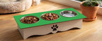 A two-bowl elevated pet feeder and waterer with a green top tray and cream-colored base, shown on a wooden floor next to a potted plant.