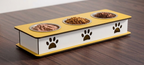 A three-compartment elevated pet feeder with a yellow top and white base, featuring metal bowls filled with wet and dry food, set on a dark wood table.