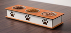 A rectangular elevated pet feeder with three metal bowls filled with dry food, placed on a dark wood tabletop. The feeder has a white front panel with three dark brown paw print cutouts and a light brown wood-grain top and base.