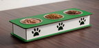 A three-compartment elevated pet feeder with a green top and white sides, featuring black paw print cutouts and metal bowls containing food, placed on a dark wood surface.
