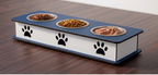 A rectangular elevated pet feeder with three bowls filled with dry and wet food, placed on a dark wood tabletop. The feeder has a white body with black paw print graphics and a dark blue top and base.