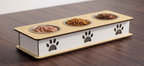 A three-compartment elevated pet feeder with a light wood top, white side panels featuring laser-cut paw prints, and stainless steel bowls filled with wet and dry food, shown on a dark wooden table.