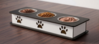 A rectangular elevated pet feeder with three stainless steel bowls on a black top and white sides, decorated with black paw prints. The feeder is shown on a dark wood table with dry and wet pet food inside the bowls.