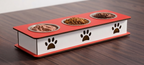A three-bowl elevated pet feeder with a red top and base, white side panels, and black paw print cutouts, shown on a dark wood tabletop with food in the bowls.