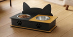 A black and yellow cat feeder and waterer with cat-ear silhouette backrest, shown on a wooden floor with food and water bowls.