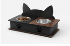 A black cat-shaped elevated pet feeder with two bowls, one filled with dry food. The feeder has a matte black body with a wood-grain top panel and base, featuring a cat head silhouette on the back, paw prints on the front, and a clean white background.