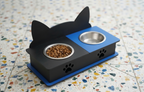 A black and blue elevated pet feeder with cat-ear silhouette sides, two stainless steel bowls, and paw-print details, shown on a speckled tile floor with food in one bowl.