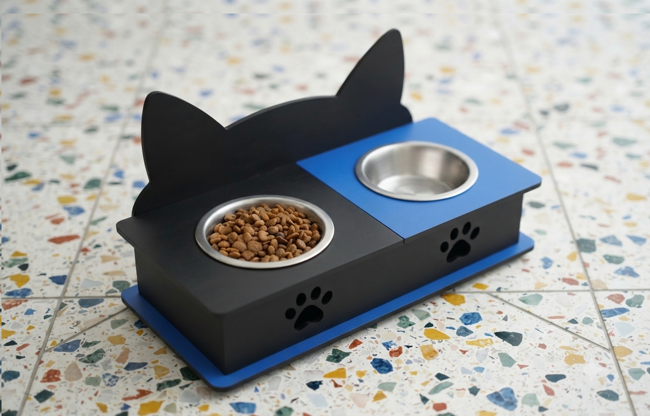 A black and blue elevated pet feeder with cat-ear silhouette sides, two stainless steel bowls, and paw-print details, shown on a speckled tile floor with food in one bowl.