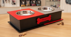 A black and red elevated pet feeder and water bowl stand with two stainless steel bowls, personalized with the name 'Mano' on a red bone-shaped plaque, set on a wooden tabletop.