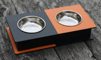 A two-compartment elevated pet feeder and water bowl stand with a matte black base, one black compartment, and one orange compartment, each holding a stainless steel bowl, shown on a weathered gray wooden surface.