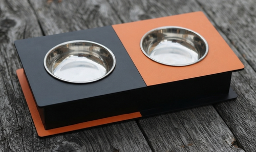 A two-compartment elevated pet feeder and water bowl stand with a matte black base, one black compartment, and one orange compartment, each holding a stainless steel bowl, shown on a weathered gray wooden surface.