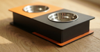 A modern two-bowl pet feeder and waterer stand with a black and orange design, featuring stainless steel bowls, shown on a light wood floor.