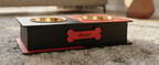 A personalized black wooden elevated pet feeder with two gold metal bowls, a red top panel, and a red bone-shaped plaque engraved with the name 'Mano', resting on a gray shag rug.