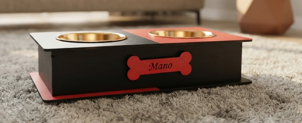 A personalized black wooden elevated pet feeder with two gold metal bowls, a red top panel, and a red bone-shaped plaque engraved with the name 'Mano', resting on a gray shag rug.
