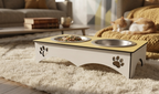 A two-bowl elevated pet feeder and water bowl with a light wood top, white base, and paw-print cutouts, shown on a shag rug in a living room with a cat sleeping on a couch in the background.