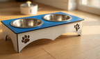 A two-bowl elevated pet feeder and water bowl stand with a blue top and white base, featuring laser-cut paw print details, shown on a light wood floor.