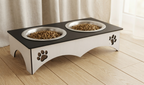A two-bowl elevated pet feeder with a black top surface and white base, featuring laser-cut paw print details and stainless steel bowls filled with dry kibble, set on a light wood floor in front of beige curtains.