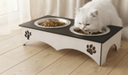 A white long-haired cat is eating from two bowls of kibble on a raised pet feeder. The feeder has a black top surface and a white base with cutout paw prints, set on a light wood floor in a bright room.
