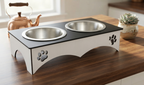 A two-bowl elevated pet feeder and waterer stand with stainless steel bowls, a black top surface, and a white base decorated with paw print cutouts, shown on a wooden table in a kitchen setting.
