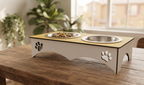 An elevated pet feeder and water bowl stand made of light wood and white material with paw print cutouts, shown on a wooden table in a bright room with a window and plants in the background.