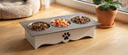 A three-bowl elevated pet feeder with a white base and gray top, shown on a wooden floor near a window with a plant and pet bed in the background.