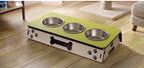 A three-bowl elevated pet feeder and waterer with a lime green top, stainless steel bowls, and a white base decorated with black paw prints and a bone graphic, shown on a wooden floor in a home setting.
