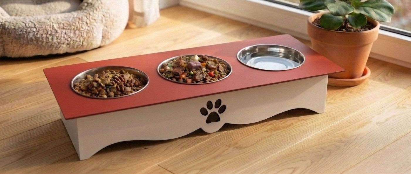 An elevated pet feeder and water bowl stand with a red top tray, three metal bowls, and a cream base with a paw print cutout, shown on a wooden floor near a window.