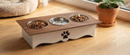 A two-tier elevated pet feeder and waterer with a dark wood top, stainless steel bowls, and a white base with a paw print cutout, set on a light wooden floor next to a potted plant.