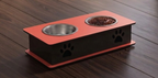 An elevated double-bowl pet feeder and water bowl stand on a dark wood floor. The stand has a black body with a coral-red top and base, featuring two stainless steel bowls (one empty, one filled with kibble) and black paw print cutouts on the front.