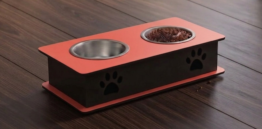 An elevated double-bowl pet feeder and water bowl stand on a dark wood floor. The stand has a black body with a coral-red top and base, featuring two stainless steel bowls (one empty, one filled with kibble) and black paw print cutouts on the front.