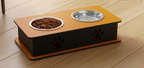 An elevated double pet feeder and waterer with a black body, orange top and base, stainless steel bowls, and paw print cutouts on a wooden floor.