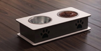 A two-bowl elevated pet feeder and water bowl stand on a dark wood floor. The stand has a white top and base with a black side panel featuring black paw print cutouts.