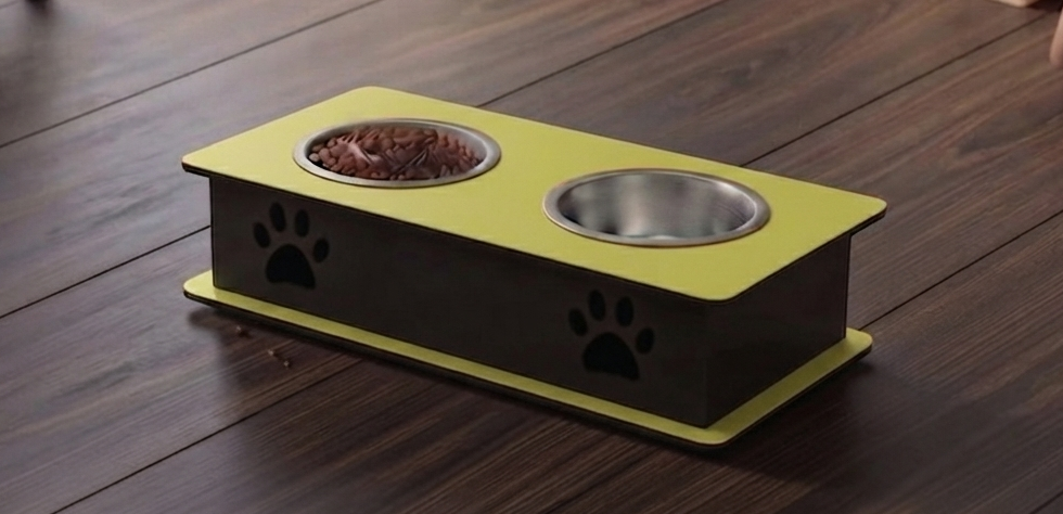 A two-bowl pet feeder and waterer elevated platform on a dark wood floor. The platform features a bright yellow top and base with black sides decorated with black paw prints, holding a bowl of dry food and an empty stainless steel bowl.