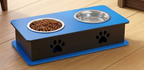 A pet elevated feeder and water bowl set on a wooden floor, featuring a blue top platform with a stainless steel food bowl filled with kibble and a stainless steel water bowl, mounted on a dark gray base with blue accents and paw print cutouts.